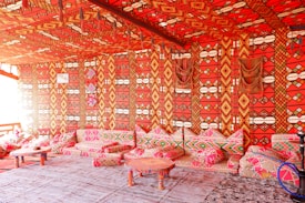 A room richly decorated with vibrant, patterned textiles covering the walls, ceiling, and cushions. The prominent colors are bright red, combined with geometric designs in orange, yellow, and black. Several low tables and cushions create a cozy seating area. The overall atmosphere suggests a traditional or cultural setting, possibly inspired by Middle Eastern or North African design.