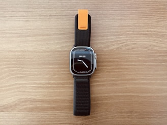 A modern smartwatch with a rectangular face and a digital display showing the time and date. The watch has a black strap with an orange accent near the top. The background is a wooden surface with a light brown color.