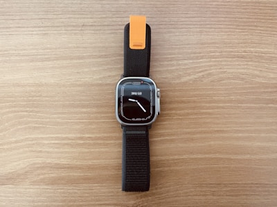 A modern smartwatch with a rectangular face and a digital display showing the time and date. The watch has a black strap with an orange accent near the top. The background is a wooden surface with a light brown color.