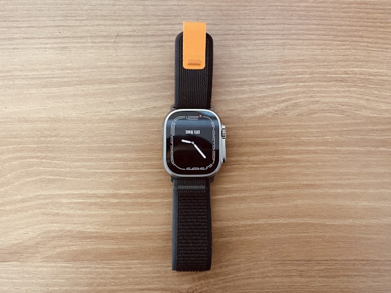 A modern smartwatch with a rectangular face and a digital display showing the time and date. The watch has a black strap with an orange accent near the top. The background is a wooden surface with a light brown color.