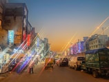 An urban street scene during dusk with numerous brightly lit signboards on both sides, indicating various hotels and shops. Vehicles, including cars and an auto-rickshaw, are visible on the road. Buildings with neon lights create a vibrant atmosphere, while a few pedestrians walk along the sidewalks.