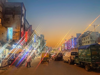 An urban street scene during dusk with numerous brightly lit signboards on both sides, indicating various hotels and shops. Vehicles, including cars and an auto-rickshaw, are visible on the road. Buildings with neon lights create a vibrant atmosphere, while a few pedestrians walk along the sidewalks.