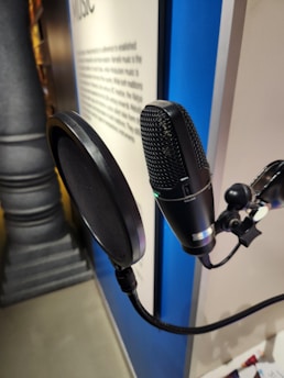 A close-up of a microphone with a pop filter mounted on a stand. The microphone is positioned in front of a blurred background that appears to be a display or sign with text. The setting suggests an indoor environment, possibly a recording studio or exhibition space.