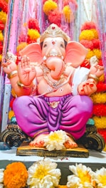 A vibrant statue of Lord Ganesha adorned with traditional ornaments and a golden crown, seated in a cross-legged position on a decorative pedestal. The statue is surrounded by colorful marigold flowers and glittering decorations in shades of red, yellow, and pink.