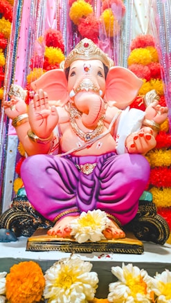 A vibrant statue of Lord Ganesha adorned with traditional ornaments and a golden crown, seated in a cross-legged position on a decorative pedestal. The statue is surrounded by colorful marigold flowers and glittering decorations in shades of red, yellow, and pink.