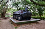 A military armored vehicle is displayed on a platform, surrounded by lush greenery and large trees. The vehicle is dark with large tires and features a rounded, robust design. It sits on a paved area edged with grass, under the canopy of towering trees that create a natural surrounding.