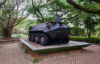 A military armored vehicle is displayed on a platform, surrounded by lush greenery and large trees. The vehicle is dark with large tires and features a rounded, robust design. It sits on a paved area edged with grass, under the canopy of towering trees that create a natural surrounding.