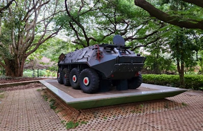 A military armored vehicle is displayed on a platform, surrounded by lush greenery and large trees. The vehicle is dark with large tires and features a rounded, robust design. It sits on a paved area edged with grass, under the canopy of towering trees that create a natural surrounding.