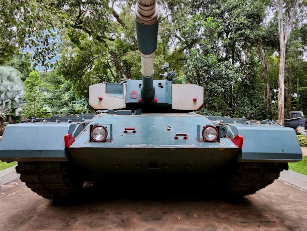 A large military tank is positioned on a concrete path surrounded by a lush, green forest. The tank is painted in shades of blue and has a large gun turret pointing forward. The surrounding trees and foliage create a dense, natural backdrop.