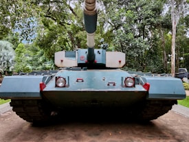 A large military tank is positioned on a concrete path surrounded by a lush, green forest. The tank is painted in shades of blue and has a large gun turret pointing forward. The surrounding trees and foliage create a dense, natural backdrop.
