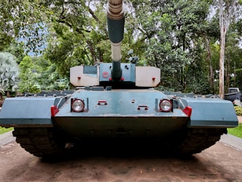 A large military tank is positioned on a concrete path surrounded by a lush, green forest. The tank is painted in shades of blue and has a large gun turret pointing forward. The surrounding trees and foliage create a dense, natural backdrop.