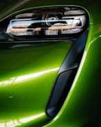A close-up view of a modern car's front headlight assembly. The headlight features sleek, black detailing with reflective surfaces and is framed by a vibrant green body panel. The smooth curves and glossy finish highlight the vehicle's futuristic and polished design.