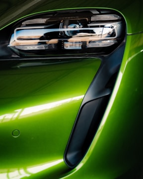 A close-up view of a modern car's front headlight assembly. The headlight features sleek, black detailing with reflective surfaces and is framed by a vibrant green body panel. The smooth curves and glossy finish highlight the vehicle's futuristic and polished design.