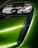 A close-up view of a modern car's front headlight assembly. The headlight features sleek, black detailing with reflective surfaces and is framed by a vibrant green body panel. The smooth curves and glossy finish highlight the vehicle's futuristic and polished design.