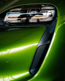 A close-up view of a modern car's front headlight assembly. The headlight features sleek, black detailing with reflective surfaces and is framed by a vibrant green body panel. The smooth curves and glossy finish highlight the vehicle's futuristic and polished design.