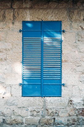 A blue shutter with horizontal slats closed against a textured stone wall. Sunlight creates dappled shadows across the surface, adding depth and contrast to the scene. The stones have a rough, varied texture, and the overall composition emphasizes the contrast between the smoothness of the shutter and the roughness of the wall.