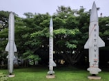 Three rocket models are displayed in a lush green outdoor area with dense trees in the background. The rocket on the left is labeled 'BrahMos,' the central rocket indicates a program by DRDO for India, and the rocket on the right is marked 'Prithvi, India.'