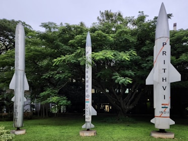 Three rocket models are displayed in a lush green outdoor area with dense trees in the background. The rocket on the left is labeled 'BrahMos,' the central rocket indicates a program by DRDO for India, and the rocket on the right is marked 'Prithvi, India.'