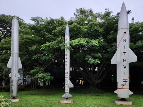 Three rocket models are displayed in a lush green outdoor area with dense trees in the background. The rocket on the left is labeled 'BrahMos,' the central rocket indicates a program by DRDO for India, and the rocket on the right is marked 'Prithvi, India.'