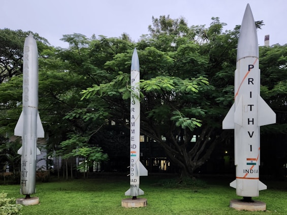 Three rocket models are displayed in a lush green outdoor area with dense trees in the background. The rocket on the left is labeled 'BrahMos,' the central rocket indicates a program by DRDO for India, and the rocket on the right is marked 'Prithvi, India.'