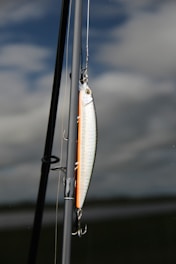 A fishing lure is attached to a fishing rod against a backdrop of cloudy sky and calm water. The lure is shiny with metallic and orange hues, featuring hooks hanging down. The rod is angled vertically with parts of the fishing line visible.