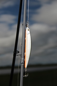A fishing lure is attached to a fishing rod against a backdrop of cloudy sky and calm water. The lure is shiny with metallic and orange hues, featuring hooks hanging down. The rod is angled vertically with parts of the fishing line visible.