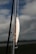 A fishing lure is attached to a fishing rod against a backdrop of cloudy sky and calm water. The lure is shiny with metallic and orange hues, featuring hooks hanging down. The rod is angled vertically with parts of the fishing line visible.