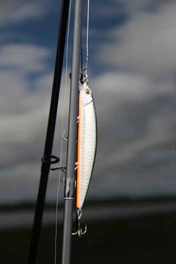 A fishing lure is attached to a fishing rod against a backdrop of cloudy sky and calm water. The lure is shiny with metallic and orange hues, featuring hooks hanging down. The rod is angled vertically with parts of the fishing line visible.