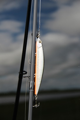 A fishing lure is attached to a fishing rod against a backdrop of cloudy sky and calm water. The lure is shiny with metallic and orange hues, featuring hooks hanging down. The rod is angled vertically with parts of the fishing line visible.