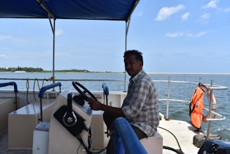 A person is seated at the helm of a boat under a blue canopy, steering the vessel on a body of water with a distant shoreline visible on the horizon. There's a life jacket hanging on the railing and some rope and equipment are observed on the boat.