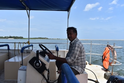 A person is seated at the helm of a boat under a blue canopy, steering the vessel on a body of water with a distant shoreline visible on the horizon. There's a life jacket hanging on the railing and some rope and equipment are observed on the boat.