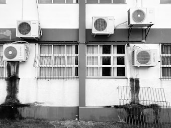 A building exterior with several air conditioning units attached to the walls. The windows have metal bars and there's noticeable black staining below some units. A metal grill is leaning against the wall near the bottom.