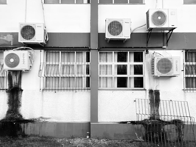 A building exterior with several air conditioning units attached to the walls. The windows have metal bars and there's noticeable black staining below some units. A metal grill is leaning against the wall near the bottom.