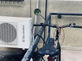 A bicycle with a black seat and handlebars is parked and locked against a metal railing. Next to it, a white air conditioning unit with the brand name 'Electrolux' is mounted on a textured wall.