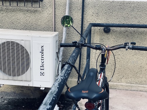 A bicycle with a black seat and handlebars is parked and locked against a metal railing. Next to it, a white air conditioning unit with the brand name 'Electrolux' is mounted on a textured wall.