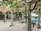 A serene outdoor space featuring a pergola with wooden beams and leafy vines overhead. Stone pillars support the structure, and there are various potted and planted greenery throughout the area. In the background, a blue swimming pool is partially visible next to a patio area with a white canopy.
