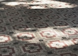 A detailed pattern of geometric tiles covers the surface, featuring a symmetrical design with red and cream-colored elements. The intricate pattern creates a feeling of depth and texture, enhanced by the play of light and shadow across the tiles.