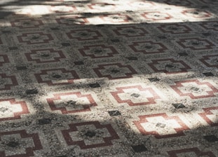 A detailed pattern of geometric tiles covers the surface, featuring a symmetrical design with red and cream-colored elements. The intricate pattern creates a feeling of depth and texture, enhanced by the play of light and shadow across the tiles.