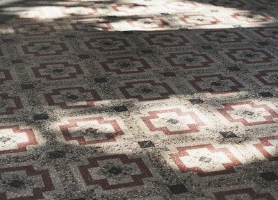 A detailed pattern of geometric tiles covers the surface, featuring a symmetrical design with red and cream-colored elements. The intricate pattern creates a feeling of depth and texture, enhanced by the play of light and shadow across the tiles.