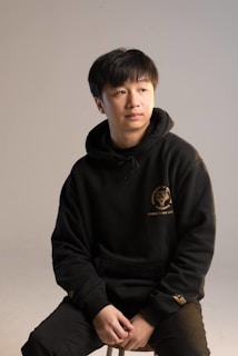 Close-up of a black hoodie with subtle golden embroidery on a minimalist beige background.