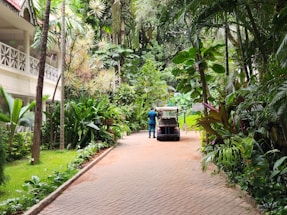A paved pathway is lined with lush tropical plants on both sides, leading to a heavily vegetated area. A person stands beside a golf cart or small utility vehicle on the path. The setting appears to be a serene garden or resort area, with a building featuring a balcony and intricate railing visible on the left side.