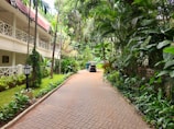 A peaceful walkway flanked by lush tropical plants and trees leads towards a two-story building with a red-tiled roof and white railings. A small vehicle, possibly a golf cart, is parked along the path, with a person standing nearby. The setting conveys a serene, resort-like environment with well-maintained greenery and a brick pathway.