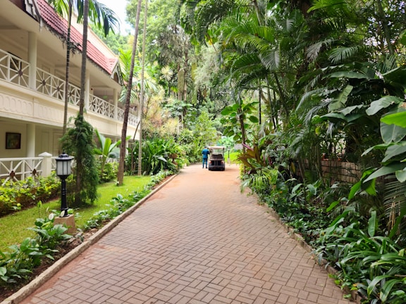 A peaceful walkway flanked by lush tropical plants and trees leads towards a two-story building with a red-tiled roof and white railings. A small vehicle, possibly a golf cart, is parked along the path, with a person standing nearby. The setting conveys a serene, resort-like environment with well-maintained greenery and a brick pathway.