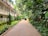 A peaceful walkway flanked by lush tropical plants and trees leads towards a two-story building with a red-tiled roof and white railings. A small vehicle, possibly a golf cart, is parked along the path, with a person standing nearby. The setting conveys a serene, resort-like environment with well-maintained greenery and a brick pathway.