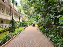 A peaceful walkway flanked by lush tropical plants and trees leads towards a two-story building with a red-tiled roof and white railings. A small vehicle, possibly a golf cart, is parked along the path, with a person standing nearby. The setting conveys a serene, resort-like environment with well-maintained greenery and a brick pathway.