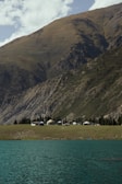 A tranquil scene featuring a series of yurts situated on a grassy plain with a backdrop of steep, rugged mountains. The foreground includes a serene body of turquoise water under a partly cloudy sky. Sparse trees line the area, complementing the remote and peaceful setting.