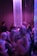 A dimly lit environment with a group of people engaging in a lively social gathering. The lighting casts a purple and blue hue over the scene with shimmering vertical strings hanging from the ceiling, creating a dynamic and energetic atmosphere.
