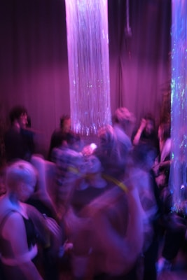 A dimly lit environment with a group of people engaging in a lively social gathering. The lighting casts a purple and blue hue over the scene with shimmering vertical strings hanging from the ceiling, creating a dynamic and energetic atmosphere.