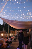 An outdoor evening event with string lights and people enjoying drinks and conversation.