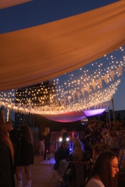 A nighttime outdoor party with string lights forming a canopy, creating a warm and festive atmosphere. People are mingling and sitting in groups under the decorative lights, with a backdrop of city buildings. A mix of colorful soft lights adds vibrancy to the scene.